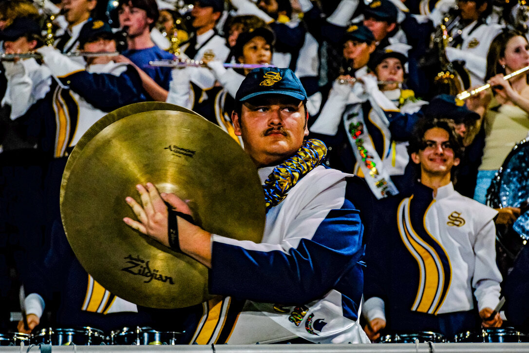 A marching band member in a blue cap holds a brass cymbal while the band plays behind him