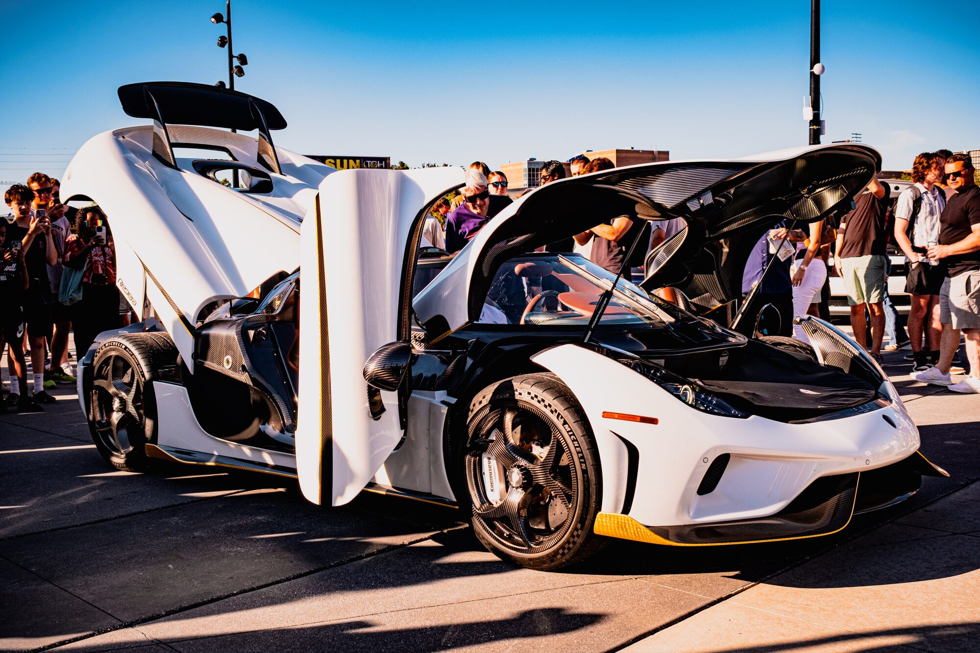 A white sports car with its doors and hood open, surrounded by people at a car show