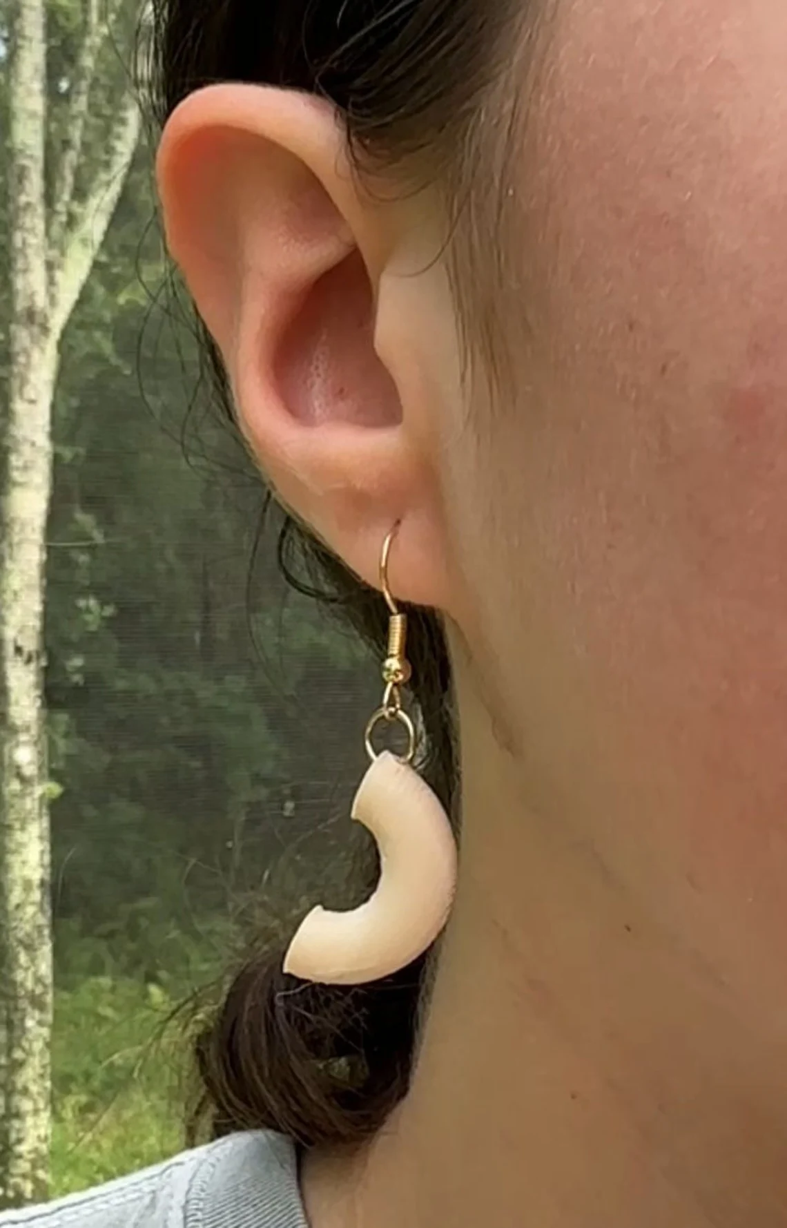A macaroni earring worn on a model's ear in profile, outdoors against green trees.